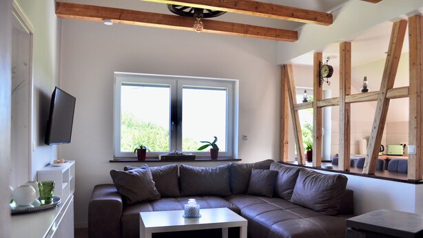 A modern living room with a brown leather sectional sofa, a white coffee table, and a TV mounted on the wall. There are two windows providing natural light, and a wooden ceiling beam is visible. - Cozy vacation home with 2 separate living units in the Thuringian Forest (Suhl)
