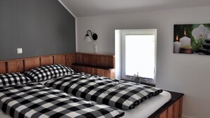 A bedroom with a wooden headboard, a large bed with checkered bedding, a bedside table, a wall-mounted lamp, and a window with a view of flowers and candles. - Cozy vacation home with 2 separate living units in the Thuringian Forest (Suhl)