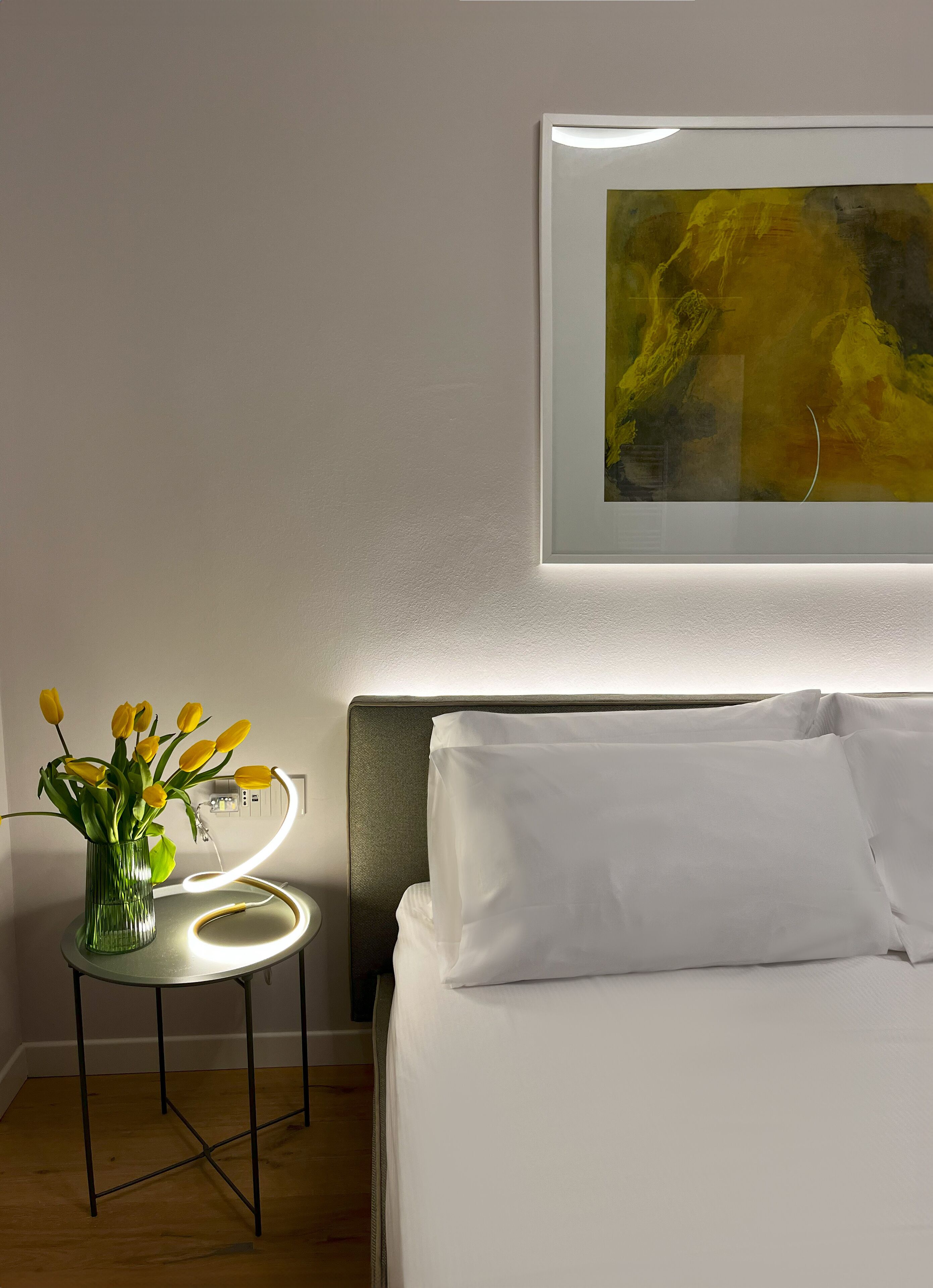 Een hotelkamer met een bed, een nachtkastje met een lamp, en een lijst met een abstract schilderij aan de muur.
