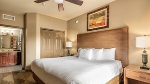 Ein Hotelzimmer mit einem großen Bett, einem Holzkopfende, einem Deckenventilator, einem Gemälde an der Wand und einem Badezimmer mit Waschtisch und Spiegel.
