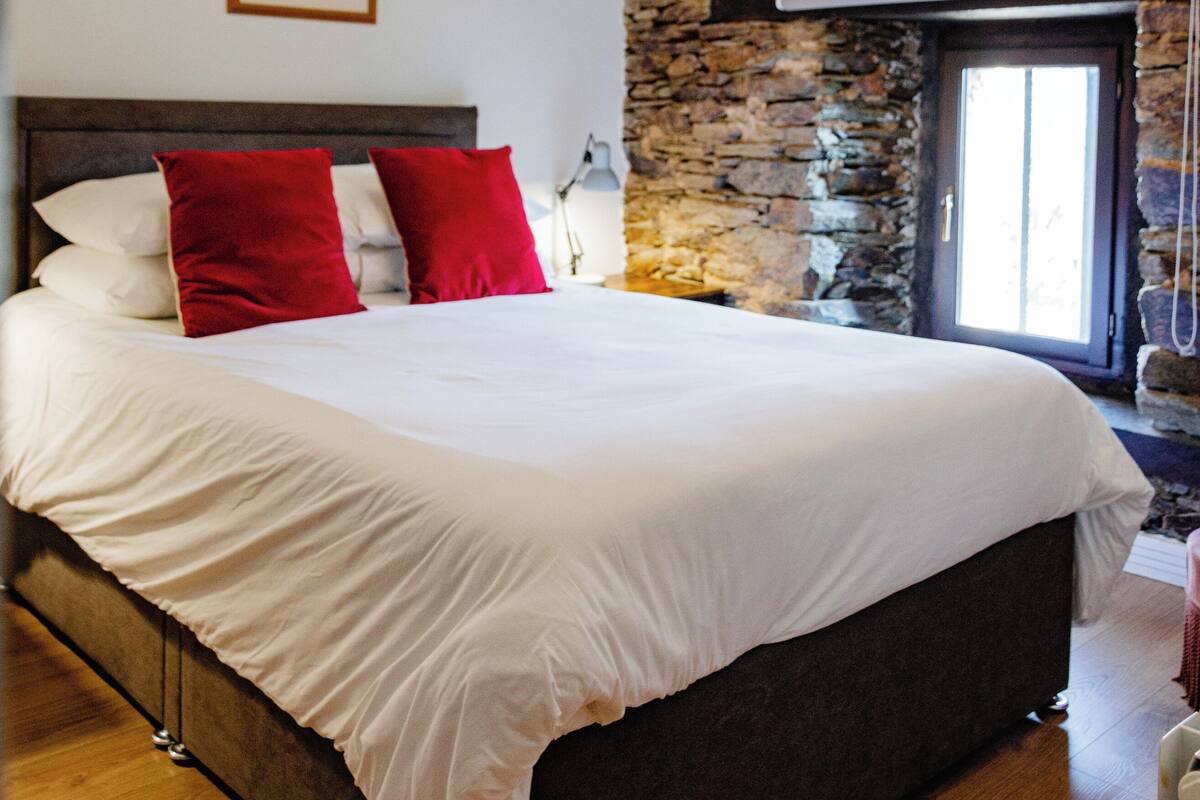 Quarto com cama grande, parede de pedra, vigas de madeira no teto e um quadro pendurado na parede.