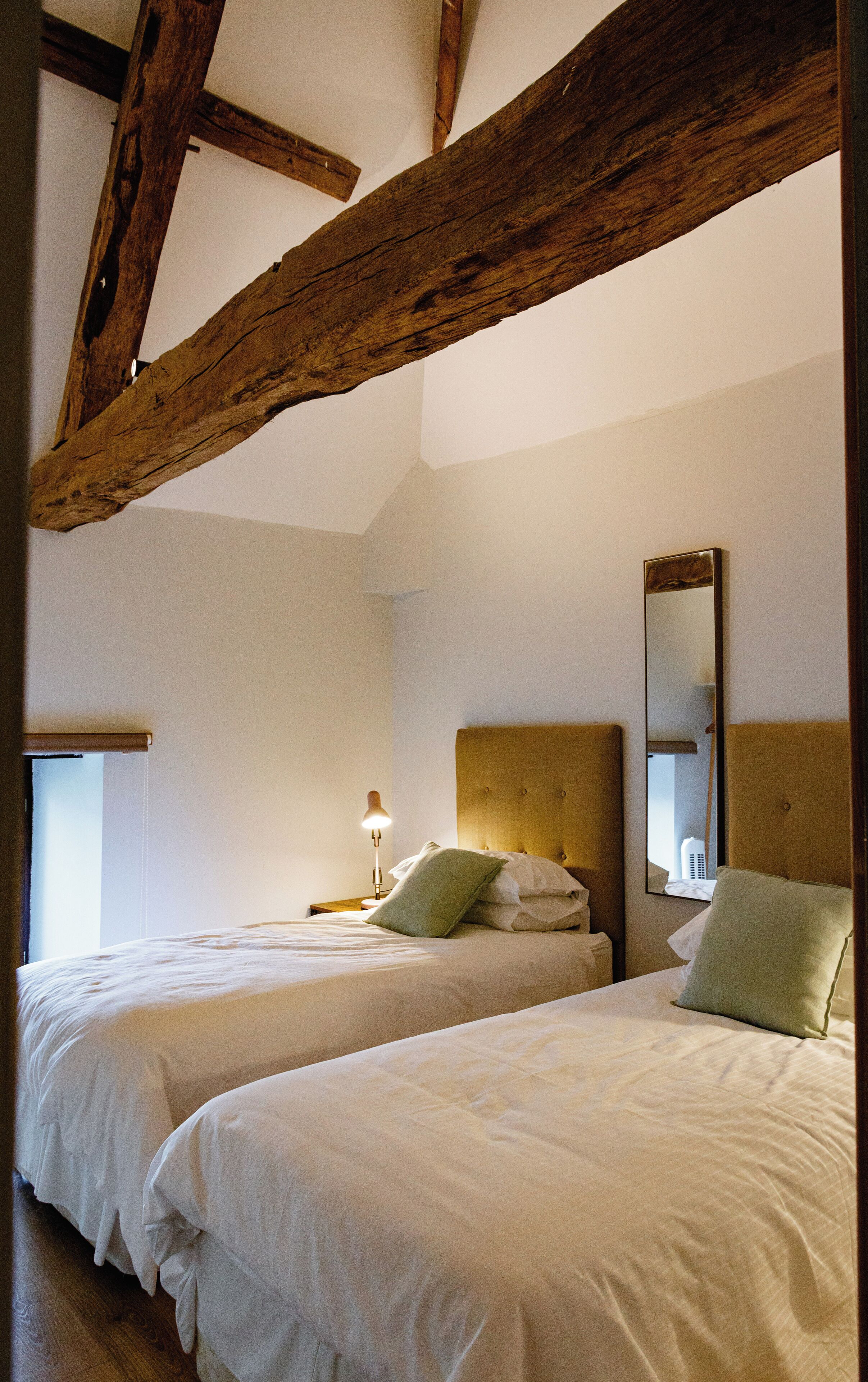 Un dormitorio con dos camas, vigas de madera, una mesita de noche con lámpara y un espejo en la pared.
