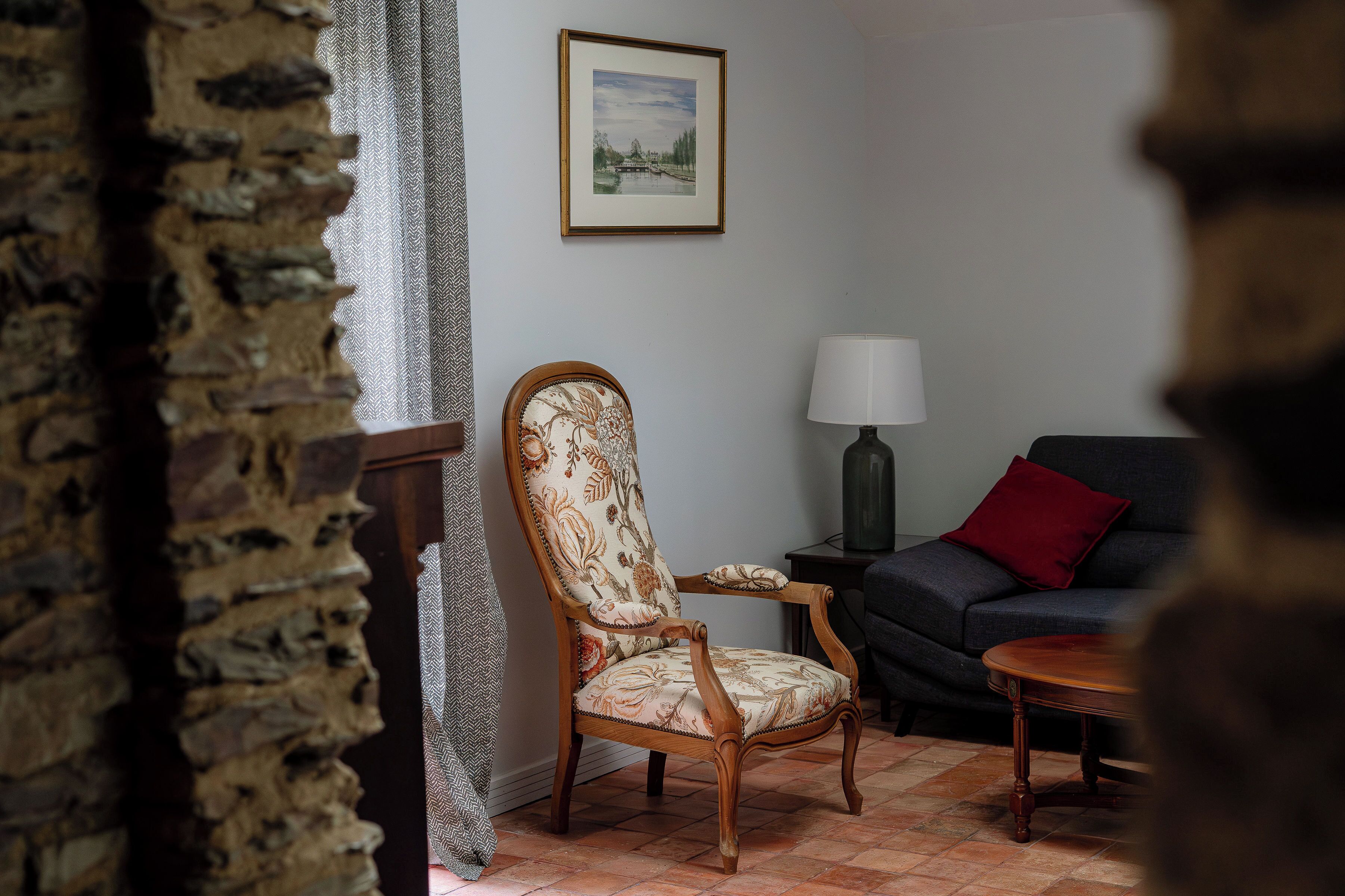 A room with a stone wall, a wooden chair with a floral cushion, a dark sofa with a red pillow, and a small table with a lamp.