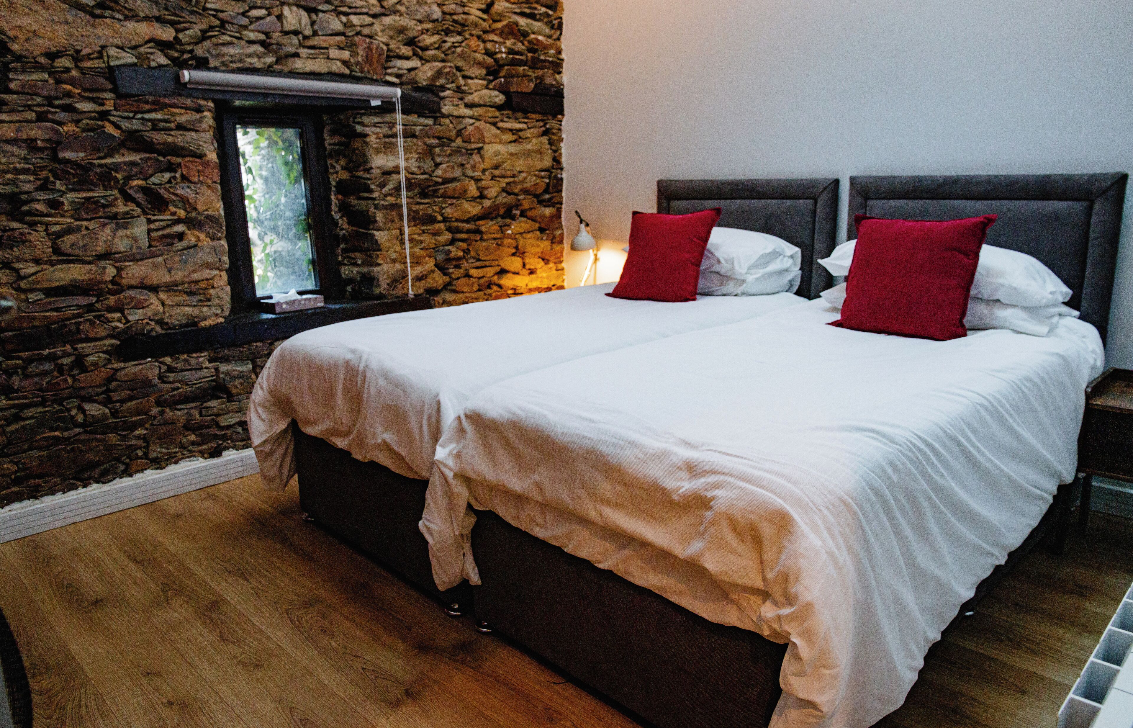 Un dormitorio con pared de piedra, una cama grande con ropa blanca y almohadas rojas, suelo de madera y una mesita de noche con lámpara.