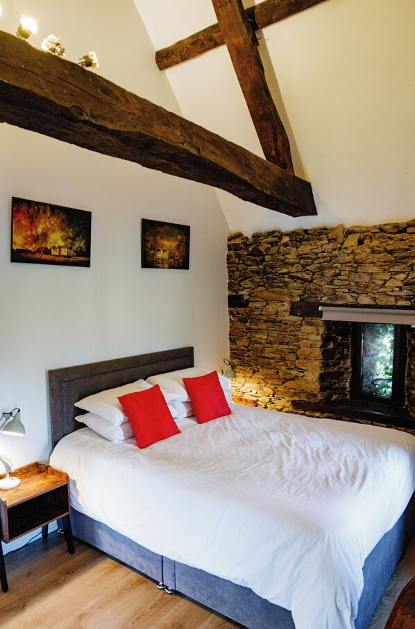 A bedroom with a stone wall, a wooden ceiling beam, a bed with white bedding and red pillows, a nightstand with a lamp, and two paintings on the wall. - Le Manoir de la Vieille Douve (Segre-en-Anjou-Bleu)