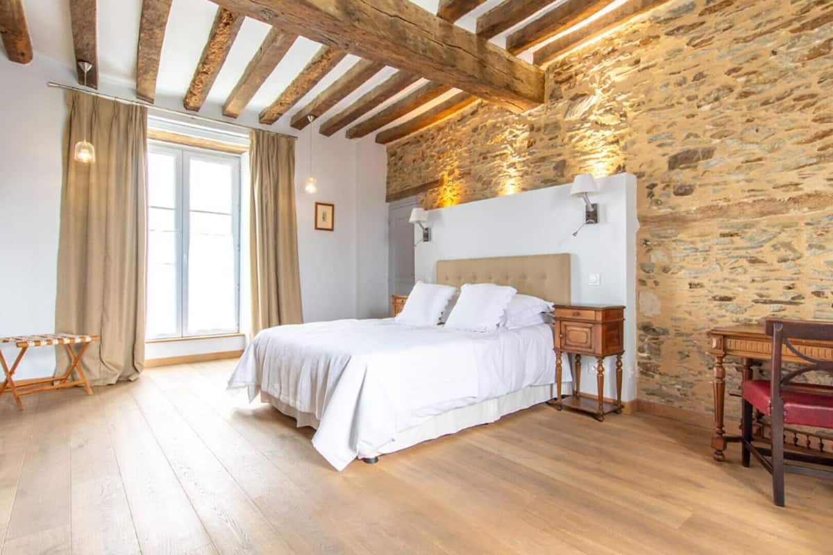 Quarto com parede de pedra, vigas de madeira, cama com roupa de cama branca, mesa de cabeceira e uma cadeira.