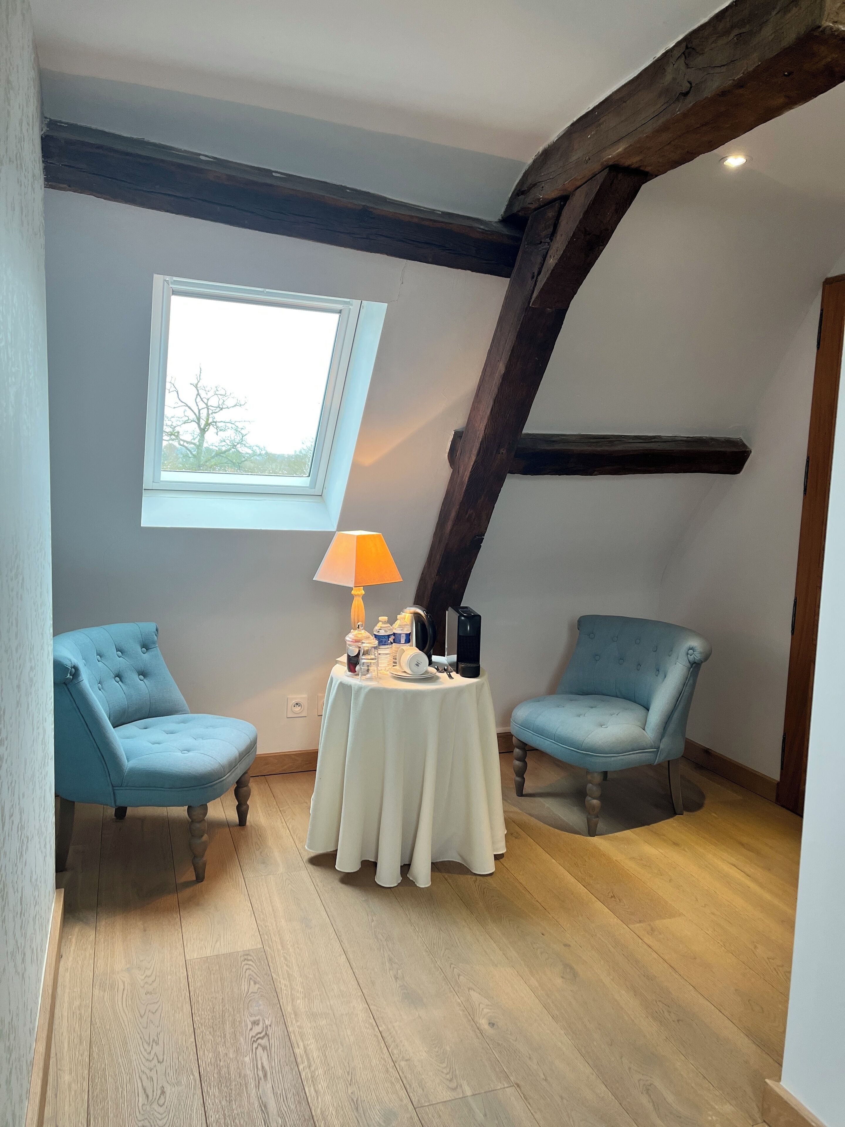 A cozy attic room with two blue armchairs, a small table with a lamp, and a window overlooking a tree.