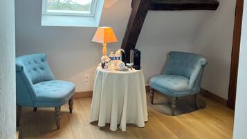 A cozy attic room with two blue armchairs, a small table with a lamp, and a window overlooking a tree.