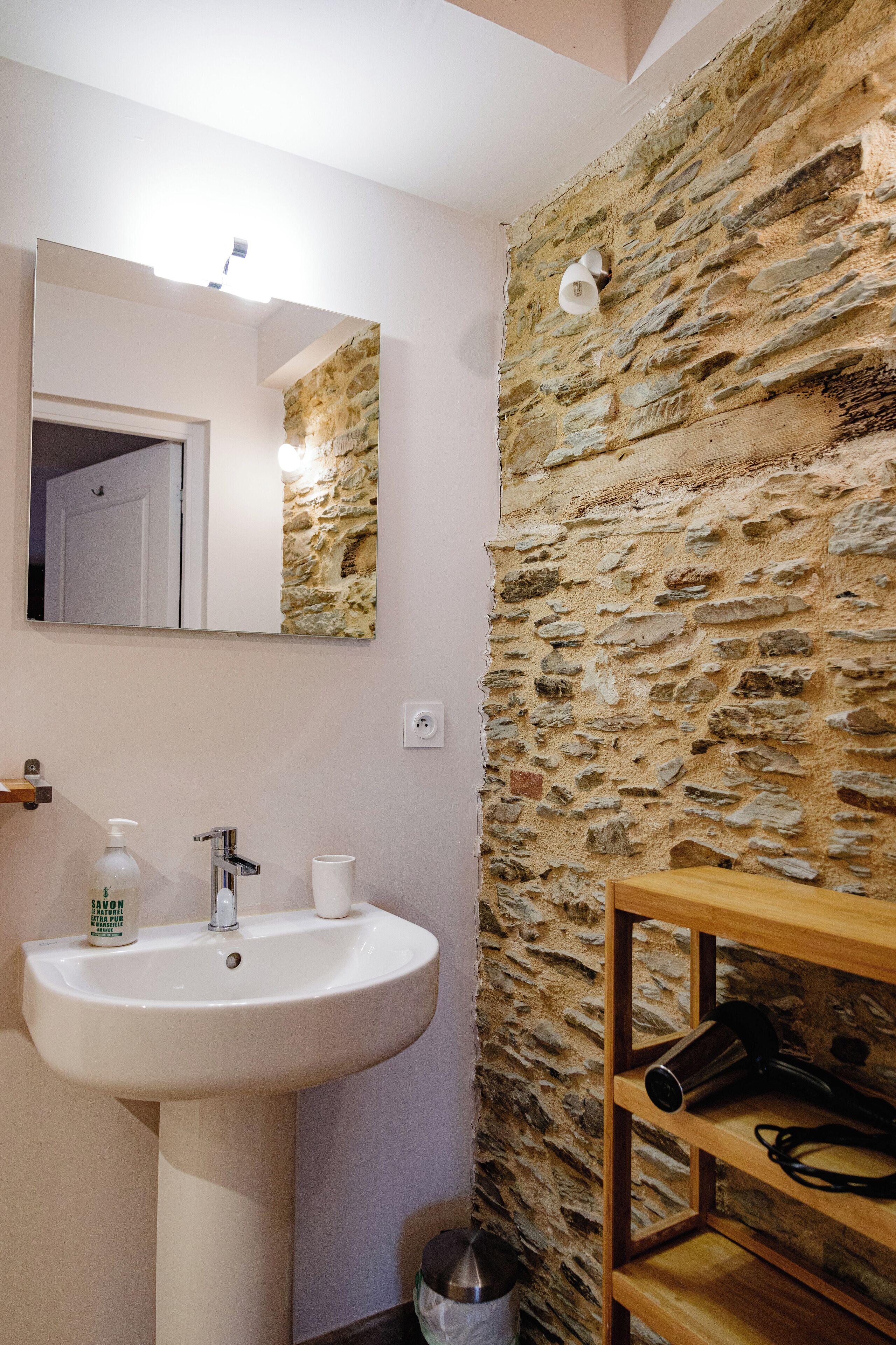 Un baño con pared de piedra, un lavabo blanco, un espejo y una estantería de madera.