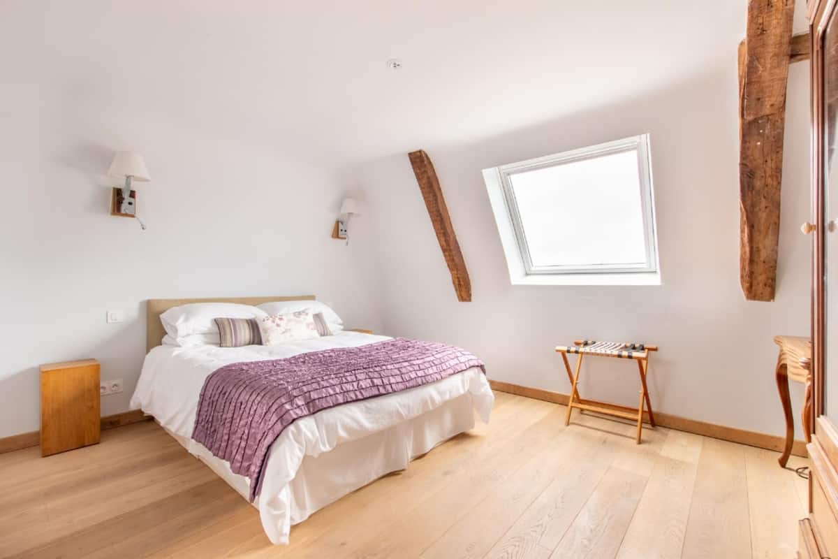 Quarto com piso de madeira, cama com roupa de cama branca e roxa, janela e cômoda de madeira.