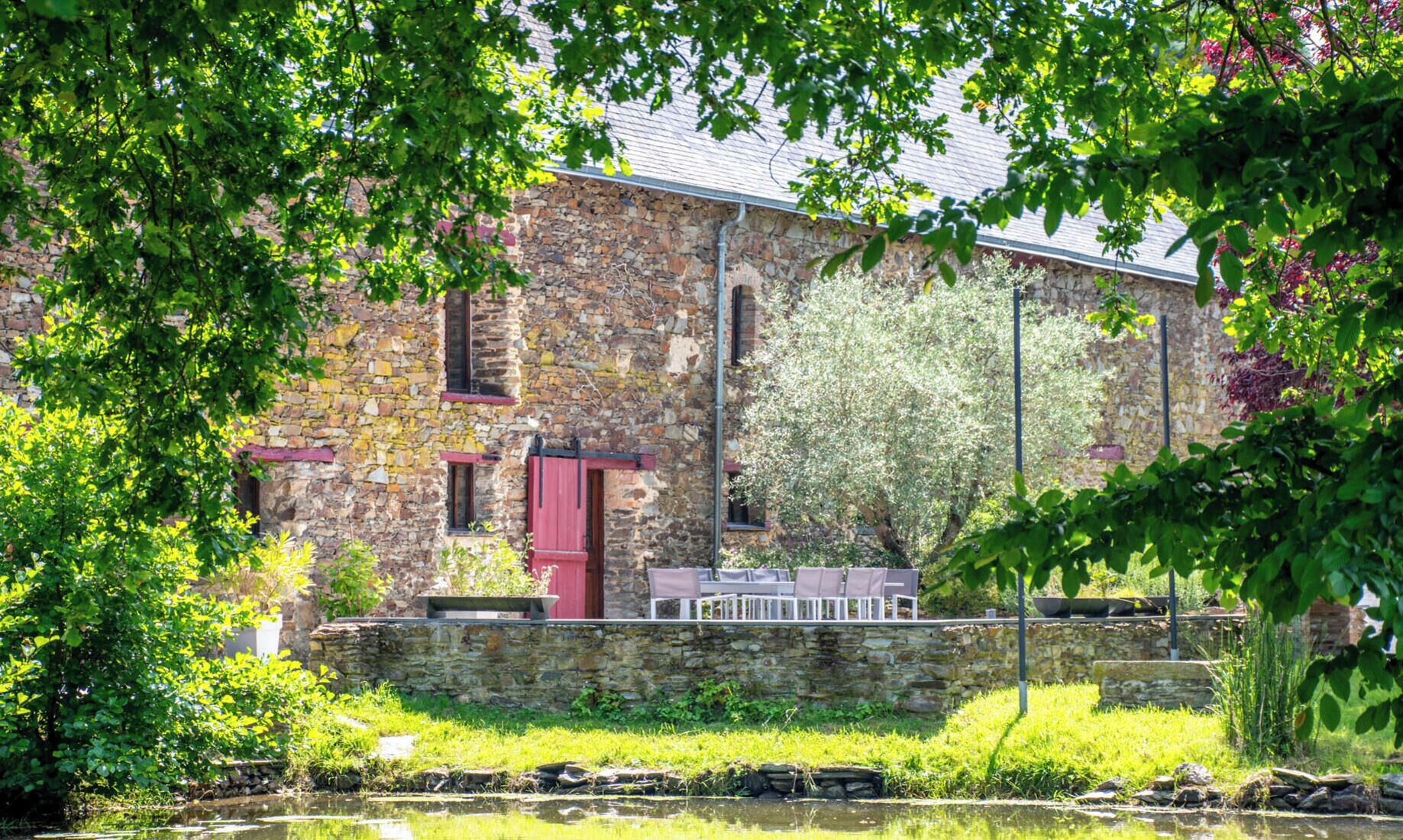 Un bâtiment en pierre avec une porte rouge, un petit balcon avec des chaises et un bassin devant.