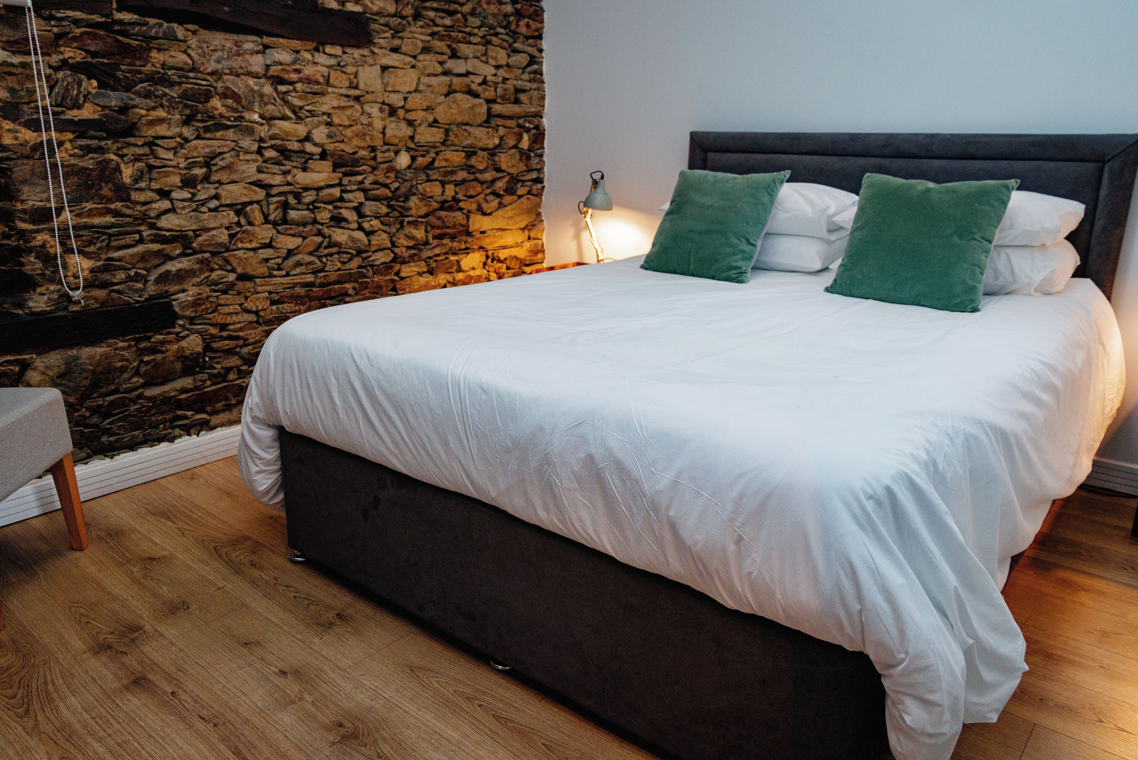 A bedroom with a stone wall, a bed with white bedding and green pillows, a bedside lamp, and a wooden floor.