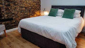A bedroom with a stone wall, a bed with white bedding and green pillows, a bedside lamp, and a wooden floor.