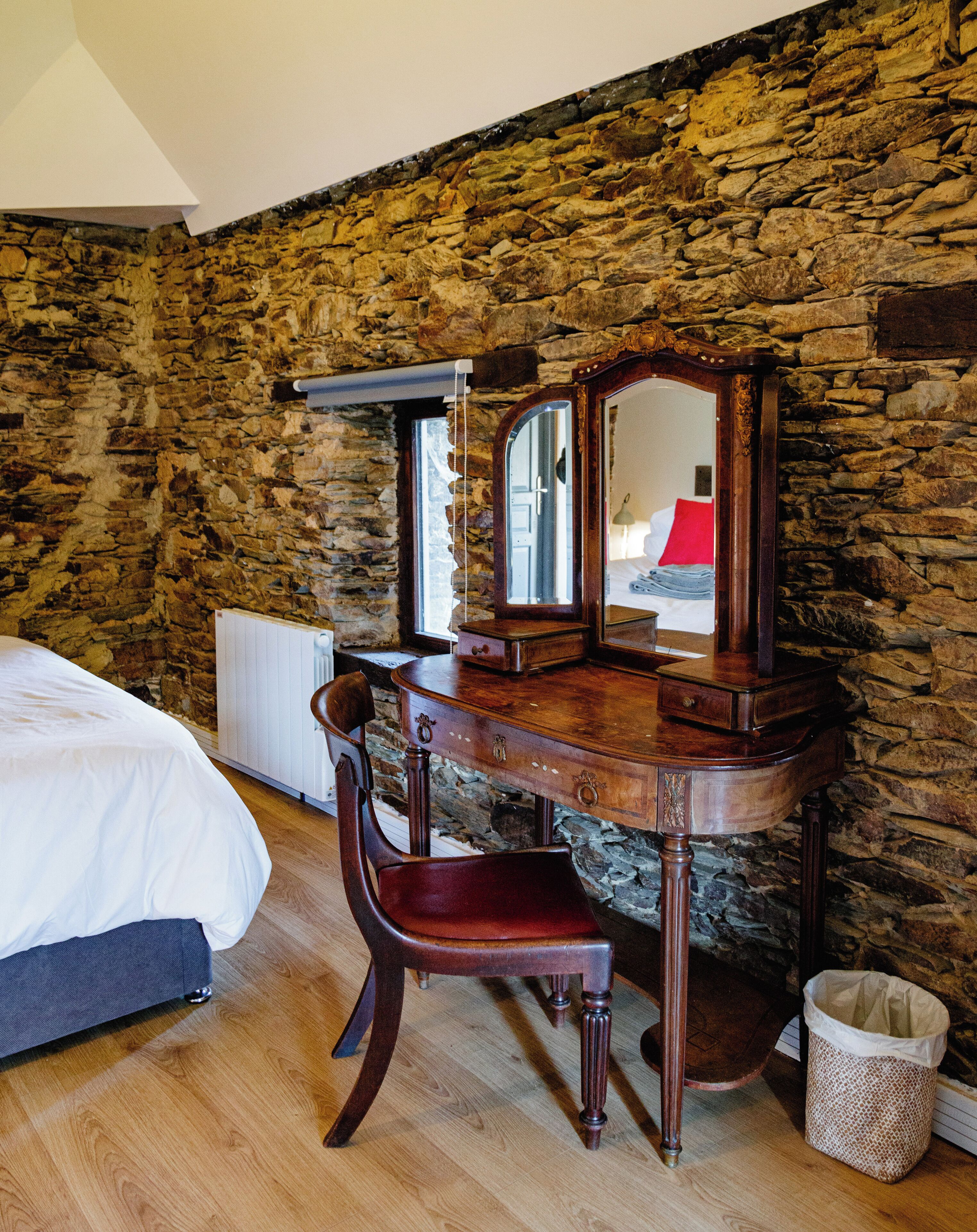 A bedroom with a wooden dresser, a mirror, a chair, a bed, and a stone wall.