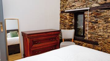 A bedroom with a wooden dresser, a chair, a bed, and a stone wall.