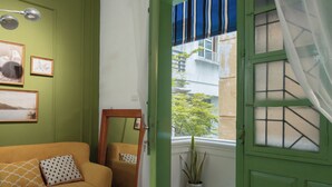 A room with a green door, a window with a view of buildings, a sofa with patterned cushions, and framed pictures on the wall. - Quiet Family 6BR Home/Large Balcony/Street View/5' to Train St/Free Laundry (Hà Nội)