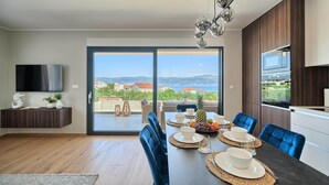 Un espace repas moderne avec une grande table dressée pour quatre personnes, un téléviseur fixé au mur et une vue sur le paysage grâce à une porte-fenêtre coulissante.