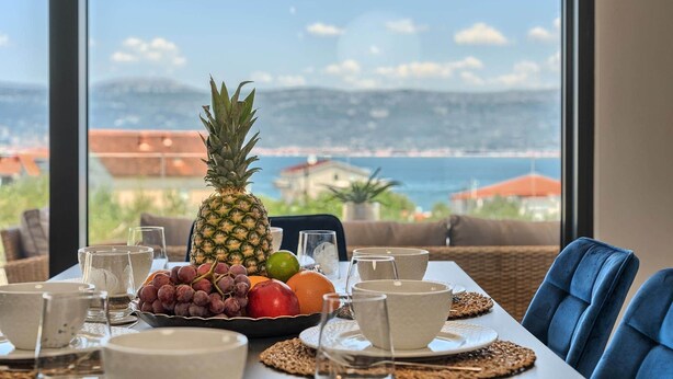 Une table dressée avec un ananas, des raisins, une pomme et un citron vert, offrant une vue panoramique sur une ville côtière à travers de grandes fenêtres.