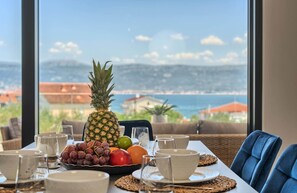 Uma mesa posta com abacaxi, uvas, uma maçã e um limão, com uma vista panorâmica de uma cidade costeira através de grandes janelas.