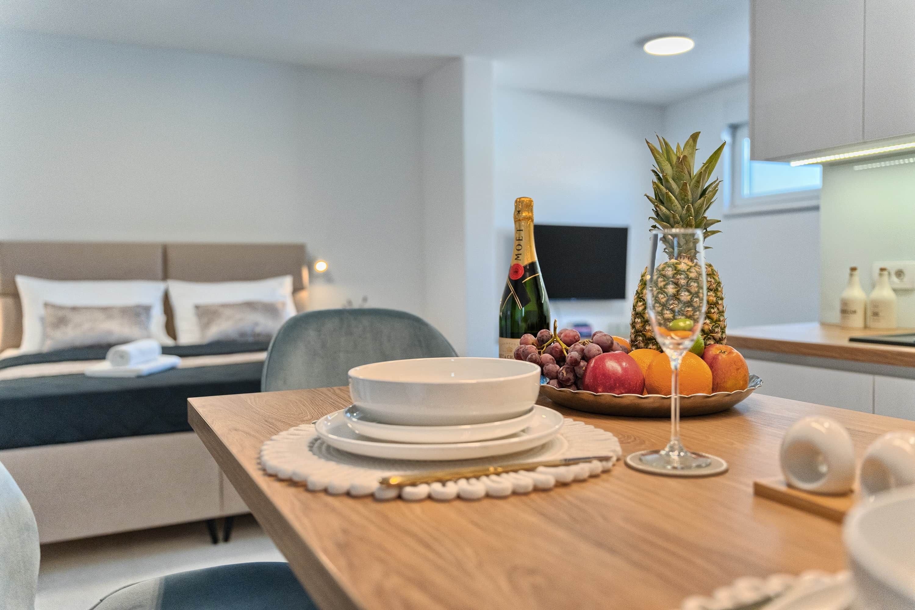 Una camera d'albergo moderna con un tavolo in legno apparecchiato con piatti, una bottiglia di champagne, un'ananas e una ciotola di frutta.