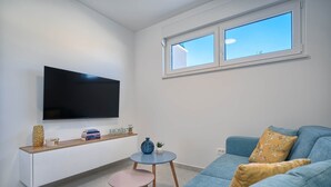 Ett modernt vardagsrum med en platt-TV som är monterad på väggen, en blå soffa, ett litet runt bord med en växt och ett fönster med utsikt över himlen.