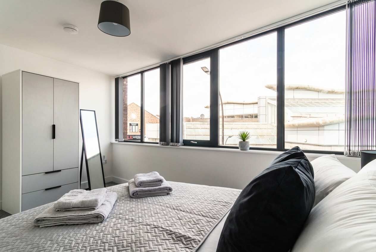 A modern bedroom with a large window, a bed with pillows and folded towels, a wardrobe, and a small plant on the windowsill.