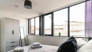 Ein modernes Schlafzimmer mit großem Fenster, einem Bett mit Kissen und gefalteten Handtüchern, einem Kleiderschrank und einer kleinen Pflanze an der Fensterbank.