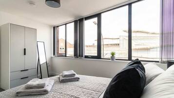 A modern bedroom with a large window, a bed with pillows and folded towels, a wardrobe, and a small plant on the windowsill.