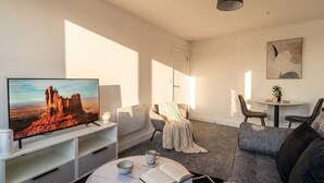 Ein modernes Wohnzimmer mit Flachbildfernseher, Couchtisch, Sofa, Sessel, einem kleinen Tisch mit Pflanze und einem Gemälde an der Wand.