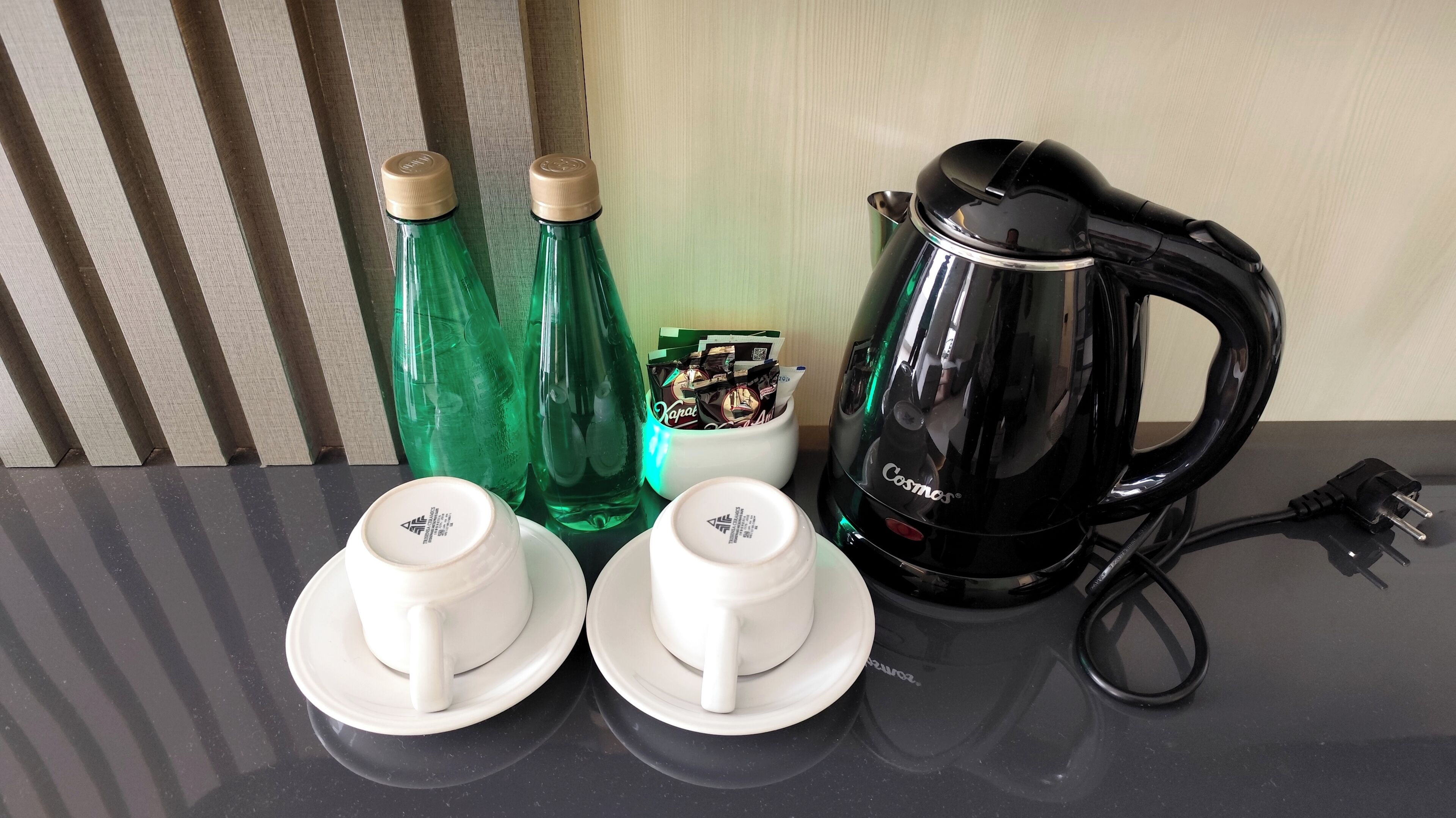 A black electric kettle, two green bottles, and two white cups with saucers on a black countertop.