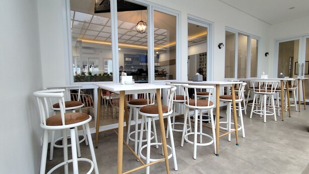 Un espace repas moderne avec des tables et des chaises blanches, de grandes fenêtres et une cuisine ouverte visible au fond.