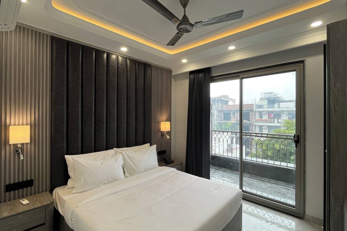 A hotel room with a bed, nightstands, a ceiling fan, a wall-mounted lamp, and a view of buildings through a balcony door.