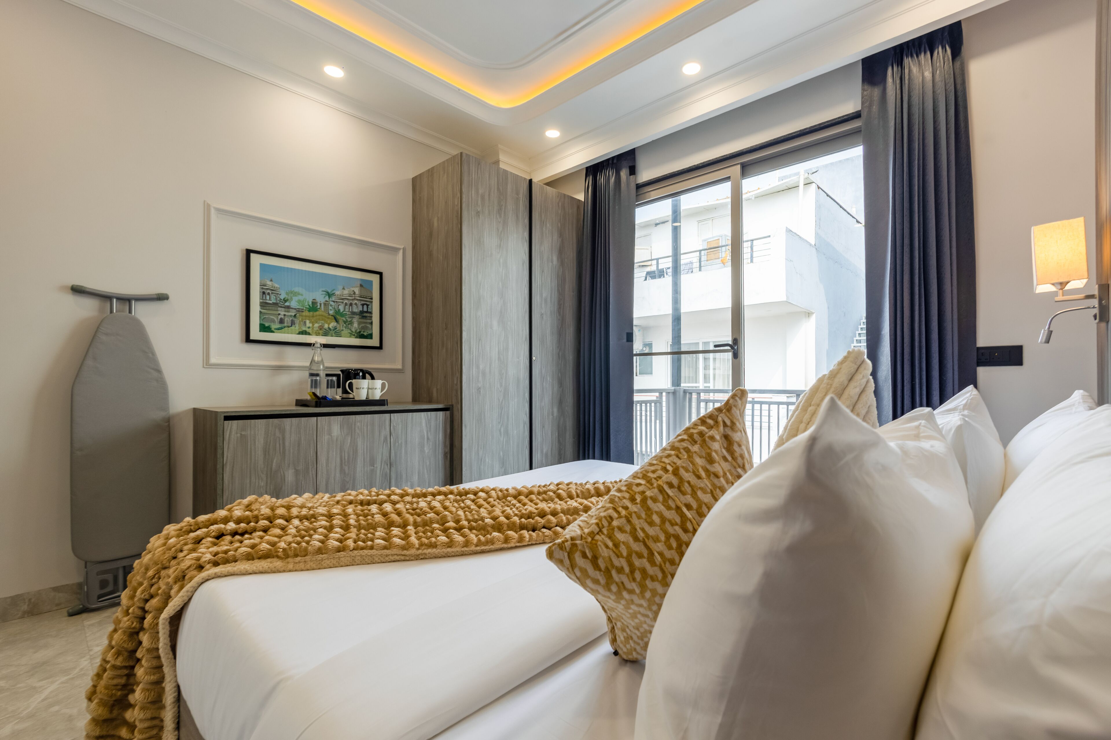 A hotel room with a bed, a desk with a coffee maker, a framed picture on the wall, and a window with curtains.