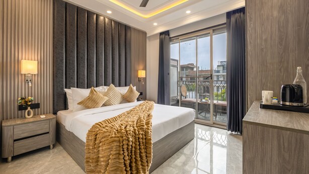 A modern hotel room with a large bed, a nightstand with a lamp, a coffee station, and a view of the city through a large window. - Saltstayz Premier Near Sector 44 Corporate Hub (Gurugram)