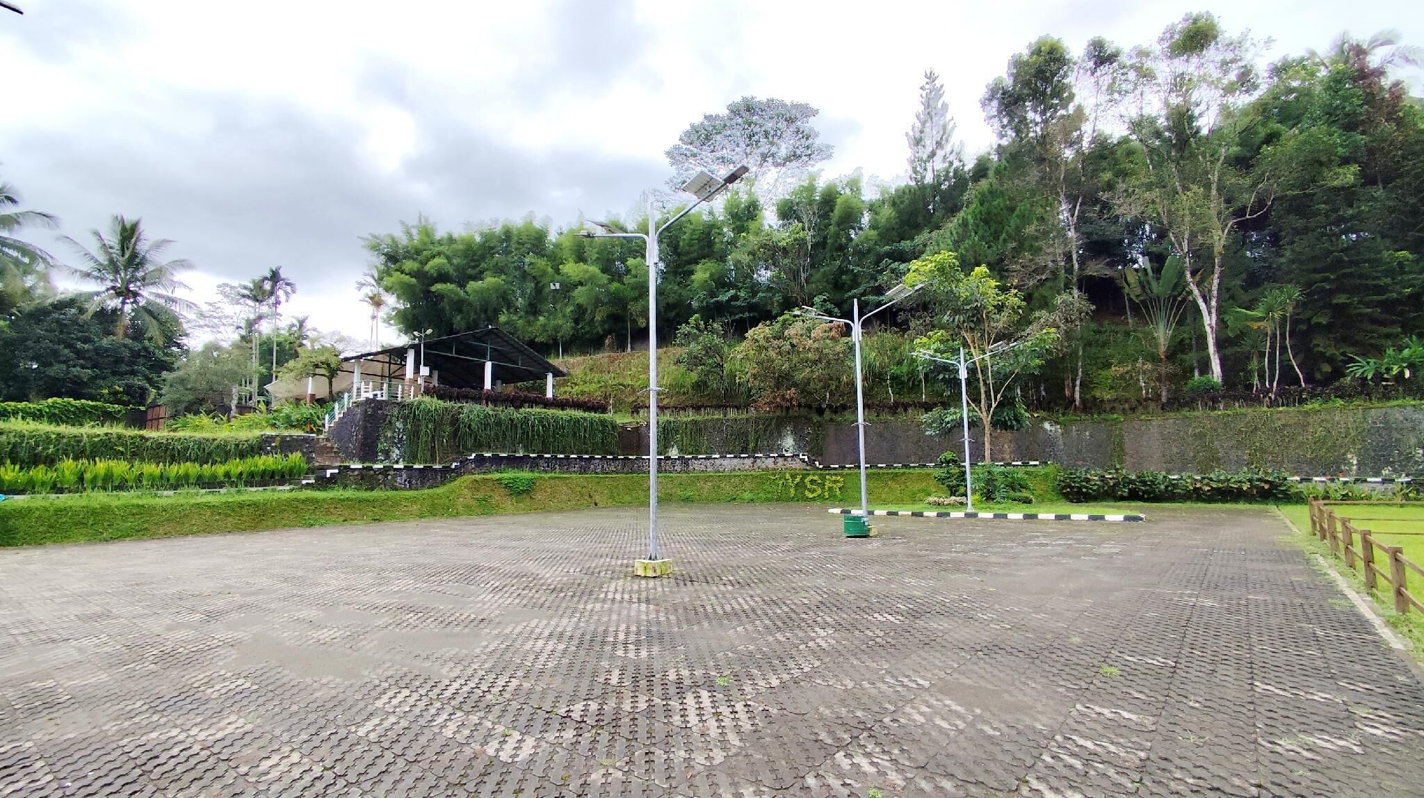 Un área pavimentada con un pabellón y farolas solares, rodeada de abundante vegetación y árboles.