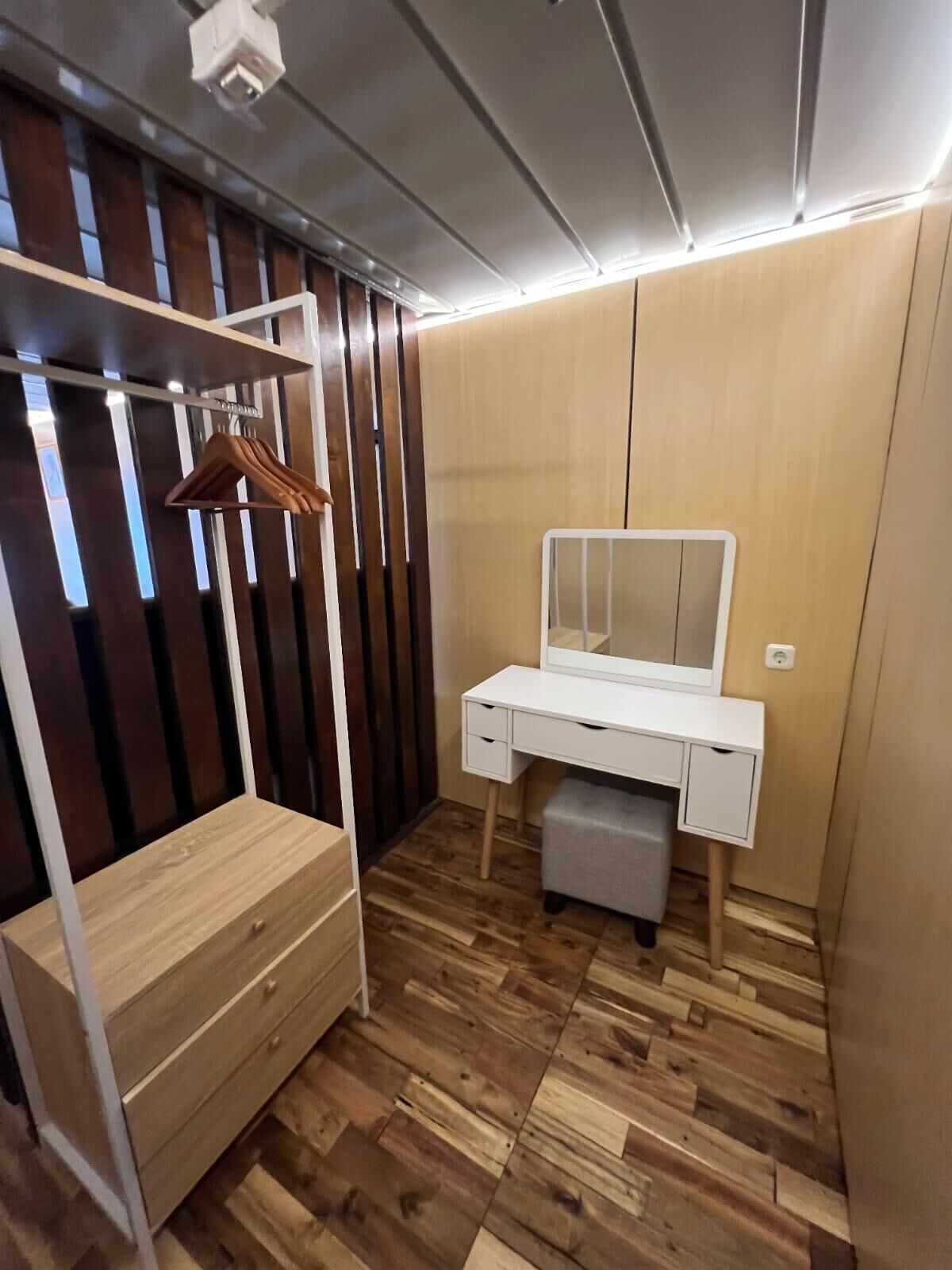 Un dormitorio compacto con un armario de madera, una mesita de noche blanca, un espejo y una percha para ropa.