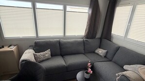 Una sala moderna con un sofá seccional gris, una mesita redonda con una flor rosa, y un comedor con una mesa y sillas blancas.