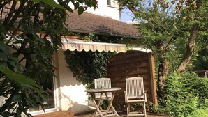 An outdoor patio with a wooden table and two chairs, surrounded by greenery and a wooden archway.