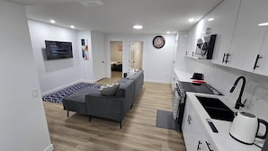A modern apartment with a grey sofa, a kitchen with white cabinets, a built-in oven, and a large clock on the wall. - Home in Chestermere Clearwater Community (Chestermere)