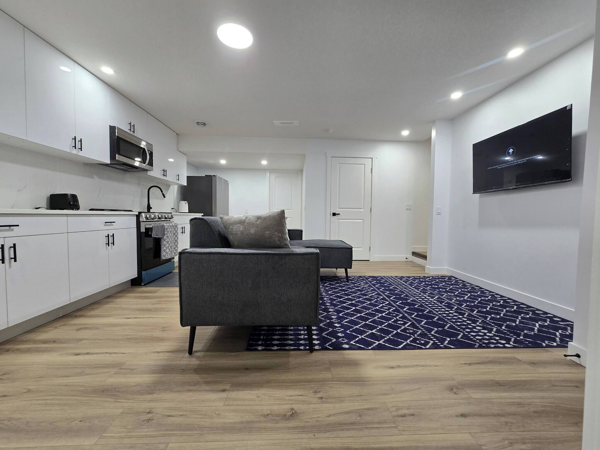A modern kitchen with white cabinets, a microwave, and a stove. There is a grey sofa with a cushion and a patterned rug on the floor. A flat-screen TV is mounted on the wall.