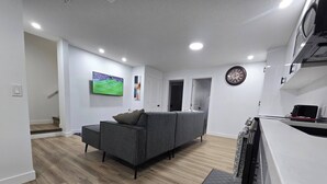A modern living room with a grey sofa, wooden flooring, a mounted TV, and a kitchen area with a microwave. - Home in Chestermere Clearwater Community (Chestermere)
