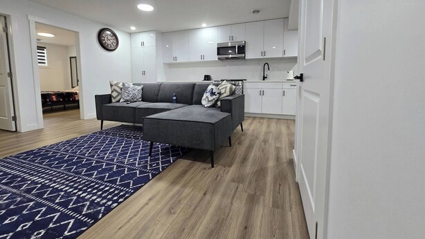 A modern living room with a grey sofa, patterned cushions, and a blue rug. A kitchen with white cabinets and a microwave is visible in the background. - Home in Chestermere Clearwater Community (Chestermere)
