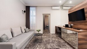 A modern hotel room with a grey sofa, a small round table with a vase, a wooden desk with a flat-screen TV, and a patterned carpet. - Sun & Snow Rakowicka Residence (Kraków)
