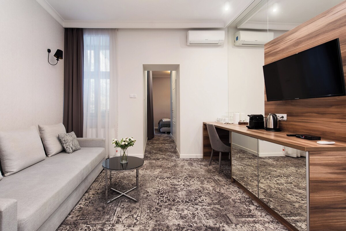 A modern hotel room with a grey sofa, a small round table with a vase, a wooden desk with a flat-screen TV, and a patterned carpet.