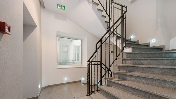 A staircase with a handrail, a window with a view, and a fire alarm on the wall. - Sun & Snow Rakowicka Residence (Kraków)