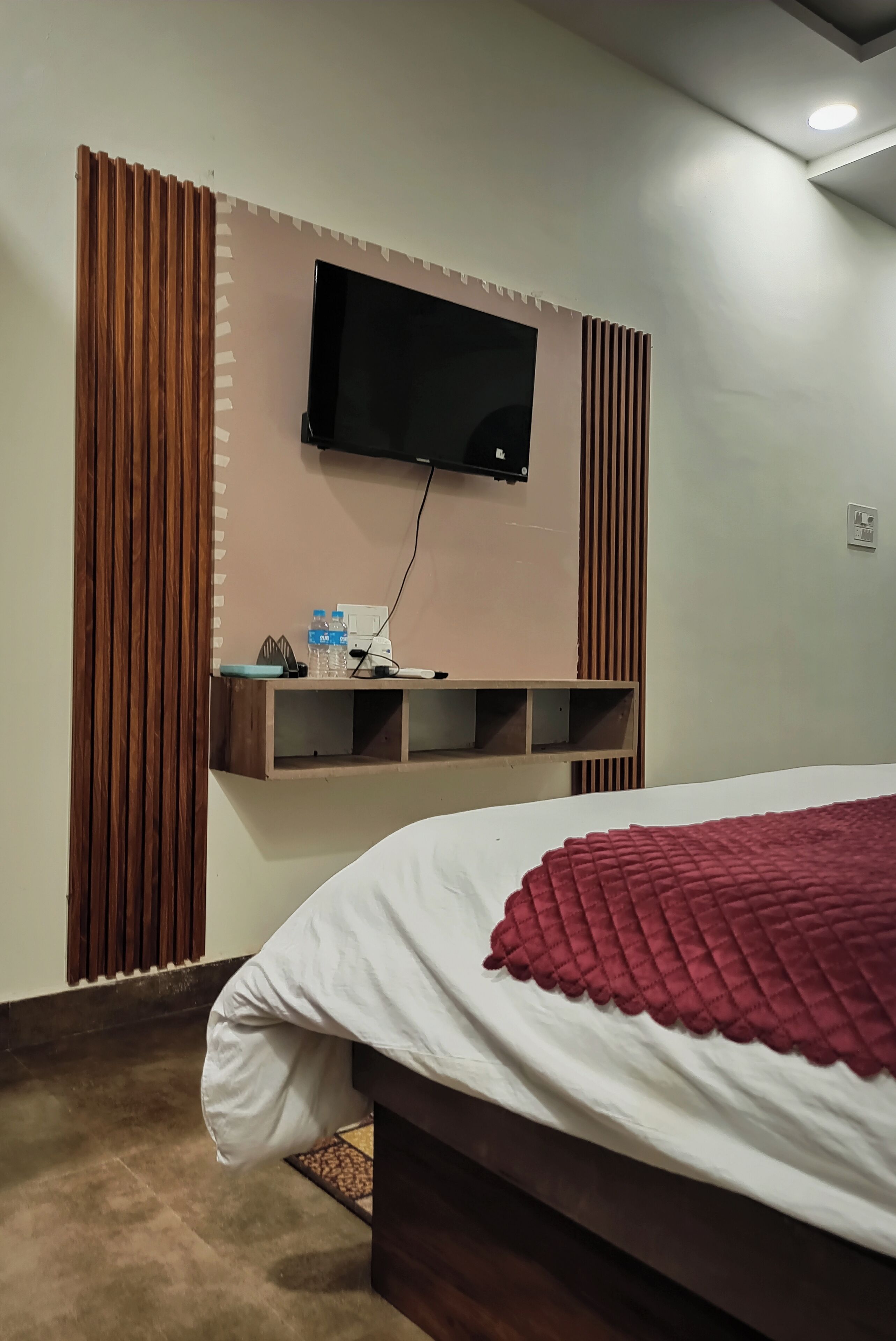 A bedroom with a bed, a TV mounted on the wall, and a shelf with water bottles.