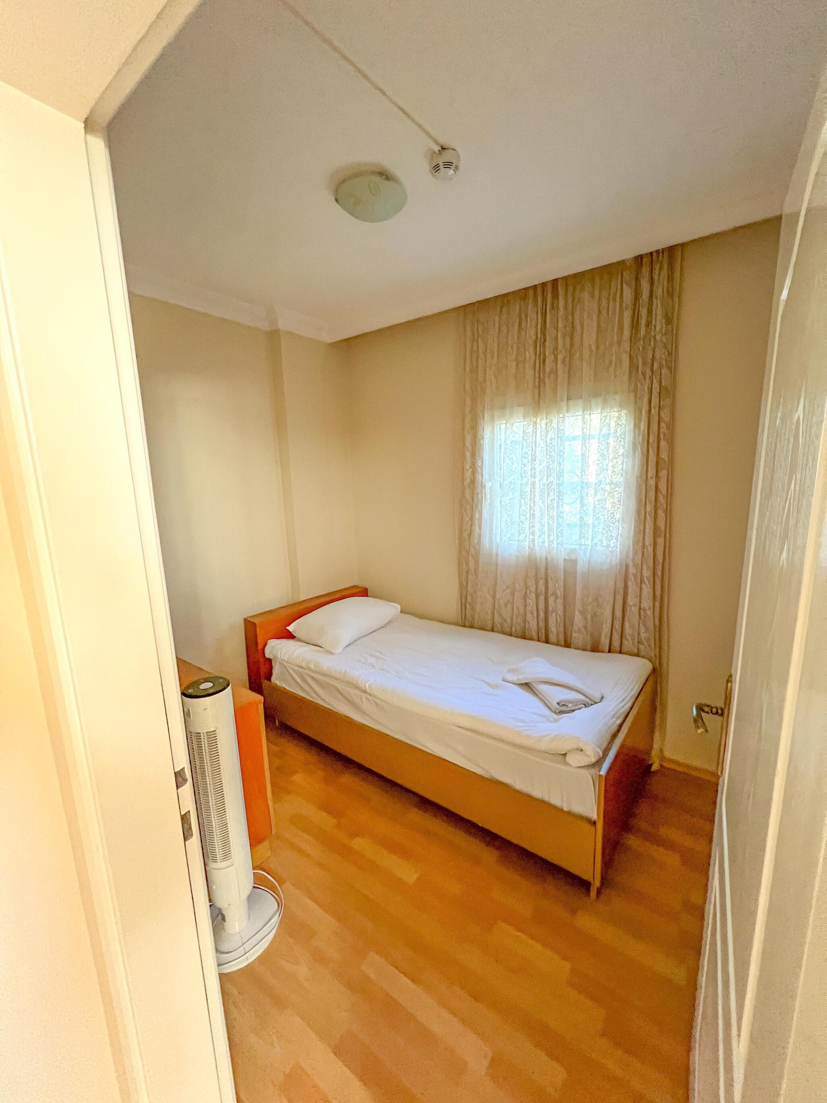 A small, single-bed room with a wooden floor, a bed with white linens, a window with curtains, and a ceiling light.