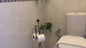 Un baño con azulejos blancos, un inodoro, un perchero para toallas con un rollo de papel higiénico y dos plantas en macetas.