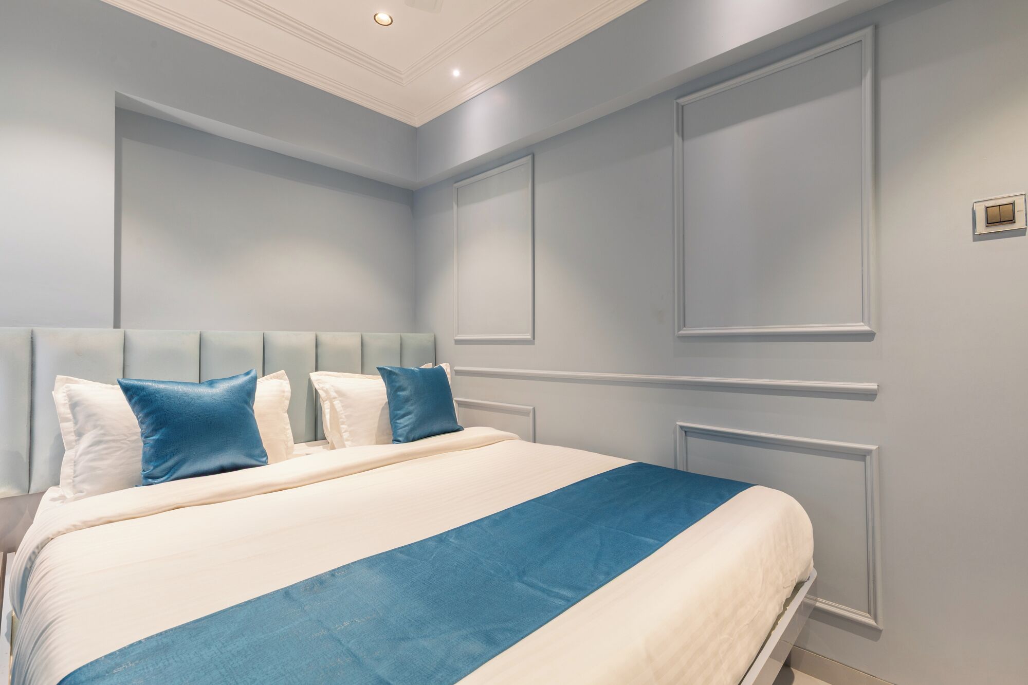 A bedroom with a large bed, a headboard with multiple panels, and a light blue wall with decorative molding.