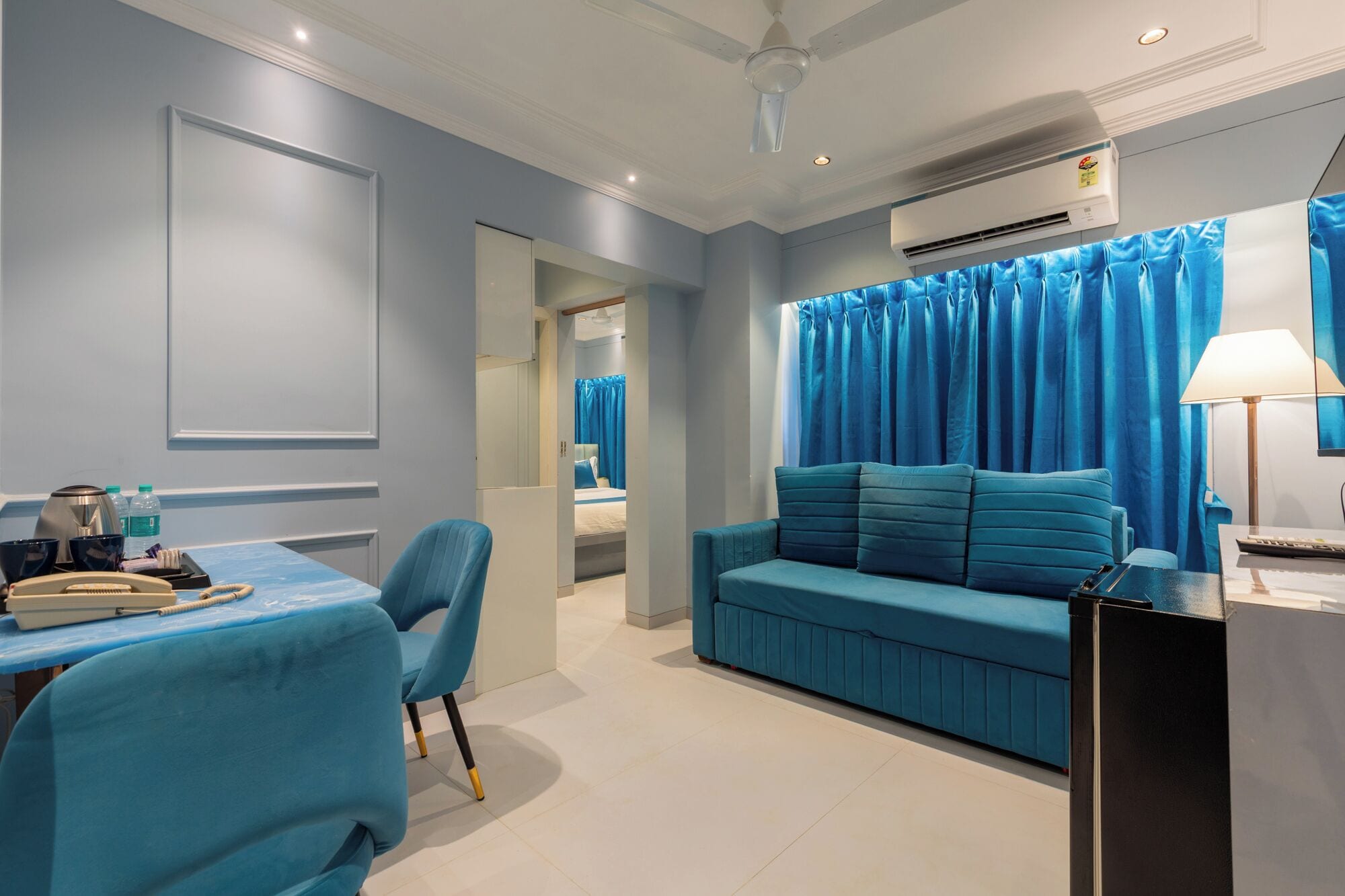 Une chambre d’hôtel moderne avec un canapé bleu, un bureau doté d’un téléphone et d’une lampe, et un appareil de climatisation fixé au mur.